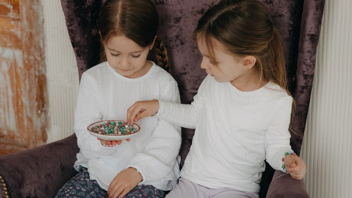 Two young girls sharing colorful beads while sitting on a purple armchair indoors.